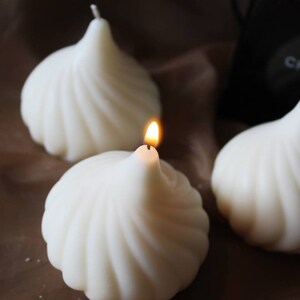 May include: Three white swirl candles, one with a lit wick. The candles are shaped like a swirl and are arranged on a brown surface.