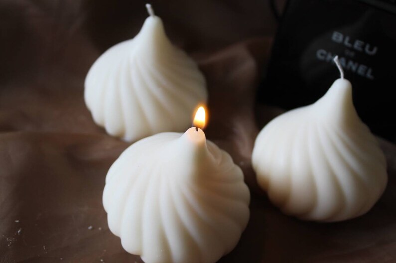 May include: Three white swirl candles, one with a lit wick. The candles are shaped like a swirl and are arranged on a brown surface.