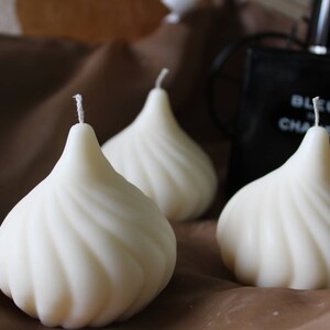 May include: Three white, swirl-shaped candles on a brown surface. The candles are shaped like a pear or a teardrop.