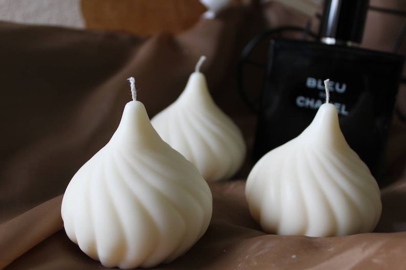 May include: Three white, swirl-shaped candles on a brown surface. The candles are shaped like a pear or a teardrop.