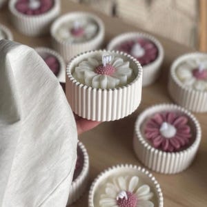 May include: White daisy-shaped candles with pink centers sit in ribbed, round, white containers. Several candles are arranged on a wooden surface. The candles have a floral design and are a decorative item.