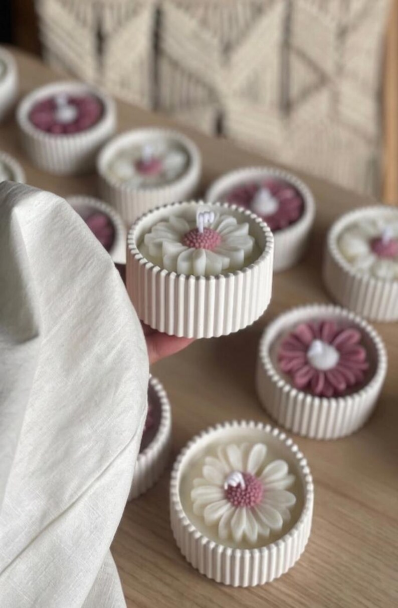 May include: White daisy-shaped candles with pink centers sit in ribbed, round, white containers. Several candles are arranged on a wooden surface. The candles have a floral design and are a decorative item.