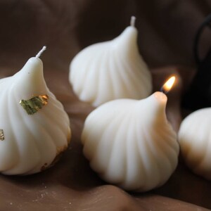 May include: Four white, decorative candles shaped like a traditional Indian sweet, with one candle lit. The candles are decorated with gold leaf.
