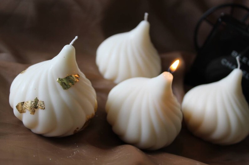 May include: Four white, decorative candles shaped like a traditional Indian sweet, with one candle lit. The candles are decorated with gold leaf.