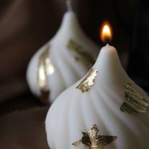 May include: Two white candles with gold leaf accents. One candle is lit and the other is unlit.