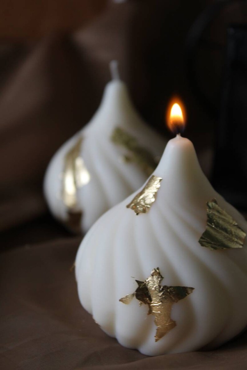 May include: Two white candles with gold leaf accents. One candle is lit and the other is unlit.