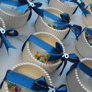 May include: White, fluted containers filled with a clear substance and colorful dried flowers. Each container is adorned with a navy blue ribbon with gold trim and a bow, and a silver "70" charm. The containers are arranged on a white surface.