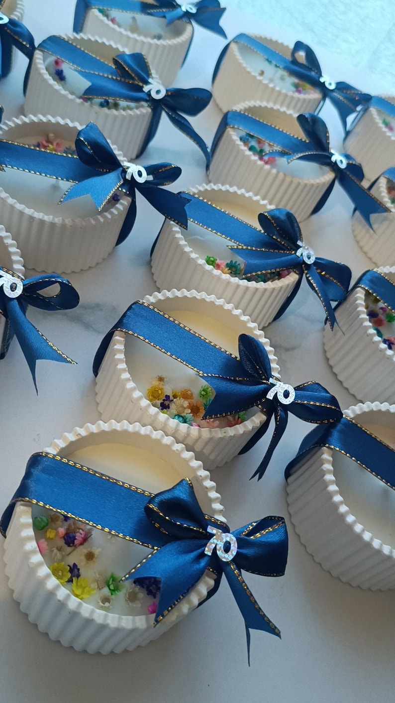 May include: White, fluted containers filled with a clear substance and colorful dried flowers. Each container is adorned with a navy blue ribbon with gold trim and a bow, and a silver "70" charm. The containers are arranged on a white surface.