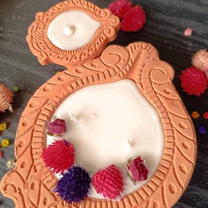 May include: A terracotta candle holder with intricate designs and a white candle. The holder is decorated with dried flowers in shades of red, pink, and purple.