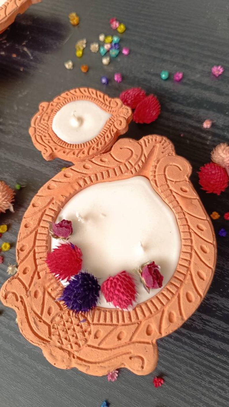 May include: A terracotta candle holder with intricate designs and a white candle. The holder is decorated with dried flowers in shades of red, pink, and purple.