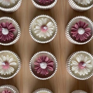 May include: A collection of daisy-shaped candles in white, and mauve, with a white wick. The candles are set in white, scalloped-edge containers, arranged on a light brown surface. The floral design adds a decorative touch.