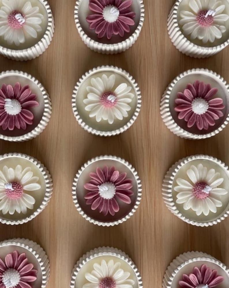 May include: A collection of daisy-shaped candles in white, and mauve, with a white wick. The candles are set in white, scalloped-edge containers, arranged on a light brown surface. The floral design adds a decorative touch.