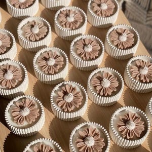 May include: A collection of round, white candles with a ribbed design, each topped with a detailed, light brown flower. The candles are arranged on a light-colored wooden surface, creating a visually appealing display.
