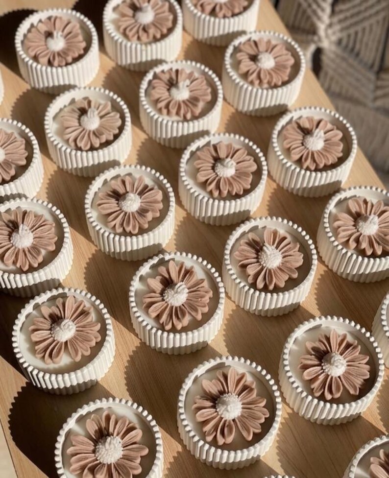 May include: A collection of round, white candles with a ribbed design, each topped with a detailed, light brown flower. The candles are arranged on a light-colored wooden surface, creating a visually appealing display.