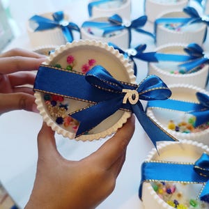 May include: White, scalloped-edge candle with colorful floral accents and a navy blue ribbon. The ribbon is tied in a bow and has a small silver "70" charm. Several similar candles are in the background.