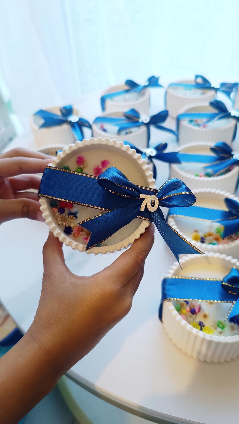 May include: White, scalloped-edge candle with colorful floral accents and a navy blue ribbon. The ribbon is tied in a bow and has a small silver "70" charm. Several similar candles are in the background.
