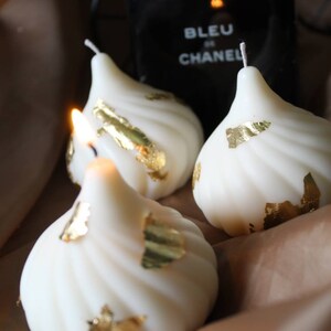 May include: Three white swirl candles with gold leaf accents. The candles are shaped like a pear or teardrop.