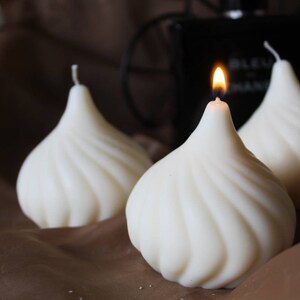 May include: Three white, swirl-shaped candles, one with a lit flame. The candles are on a brown surface.