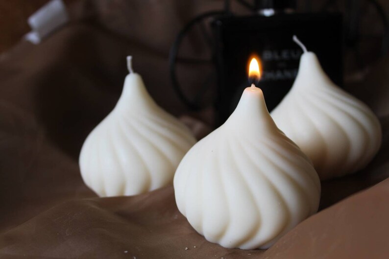 May include: Three white, swirl-shaped candles, one with a lit flame. The candles are on a brown surface.