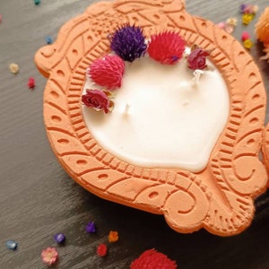 May include: A terracotta candle holder with a white candle and dried flowers in shades of pink, purple, and red. The holder has a decorative design and is on a dark wooden surface.