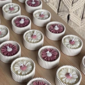 May include: A collection of daisy-shaped candles in white, pink, and burgundy. The candles are set in ribbed, round, white containers. The candles are arranged on a light brown wooden surface. The background features a macrame wall hanging.