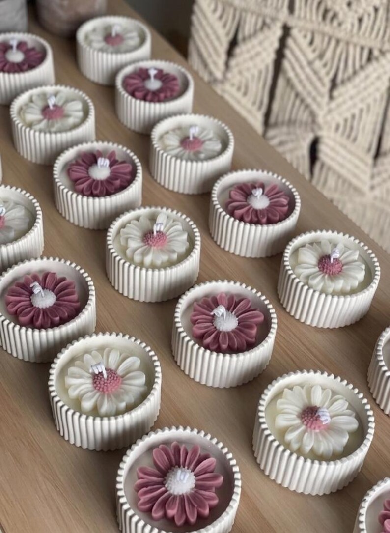 May include: A collection of daisy-shaped candles in white, pink, and burgundy. The candles are set in ribbed, round, white containers. The candles are arranged on a light brown wooden surface. The background features a macrame wall hanging.