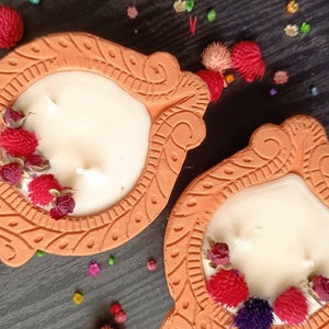 May include: Two terracotta candle holders with intricate designs. Each holder features a white candle with dried flowers and a white background. The holders are on a dark wood surface with colorful confetti.