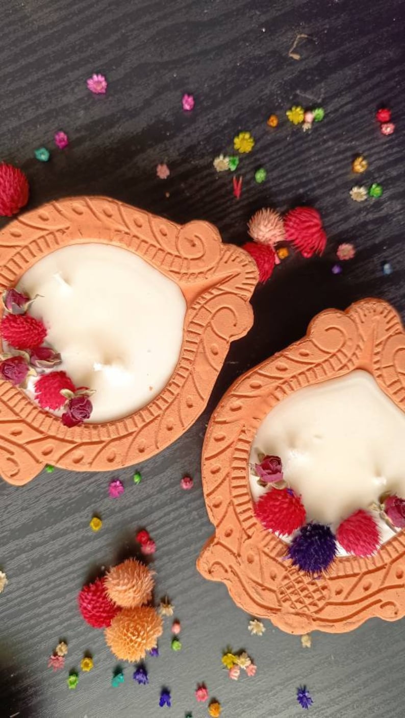 May include: Two terracotta candle holders with intricate designs. Each holder features a white candle with dried flowers and a white background. The holders are on a dark wood surface with colorful confetti.