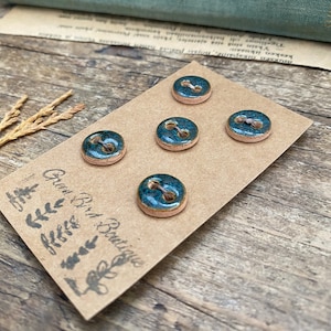 Peut inclure: Cinq boutons en céramique émaillée bleu-vert et bords bruns sont présentés sur une carte marron. Les boutons ont deux trous pour la couture. La carte est estampillée du texte "Green Bird Boutique" et de motifs de feuilles décoratives.