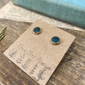May include: A pair of small, round, teal blue ceramic earrings with gold accents. The earrings are displayed on a brown card with the text "Green Bird Boutique".