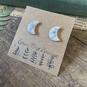 Könnte beinhalten: Ein Paar weiße Keramik-Halbmond-Ohrringe mit einer craquelé-Glasur. Die Ohrringe werden auf einer braunen Karte mit dem Text "Green Bird Boutique" und einem kleinen Blattdesign präsentiert.