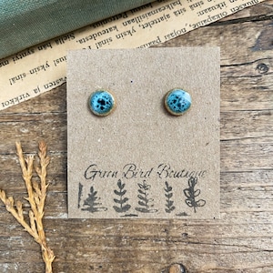 May include: A pair of round stud earrings with a turquoise speckled design and gold-colored edges. The earrings are displayed on a brown card with the text "Green Bird Boutique" and a tree illustration.