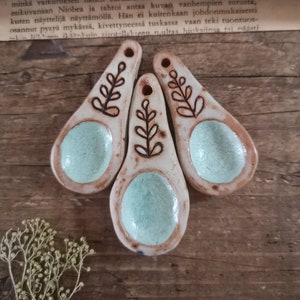 May include: Three handmade ceramic spoons with a light brown rim, a turquoise center, and a black leaf design. The spoons are arranged on a wooden surface, showcasing their unique, rustic charm.