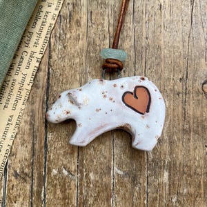 May include: A white ceramic bear pendant with brown speckles and a terracotta-colored heart detail on its side. The bear is suspended from a brown leather cord featuring a small, translucent seafoam green glass bead above the pendant.