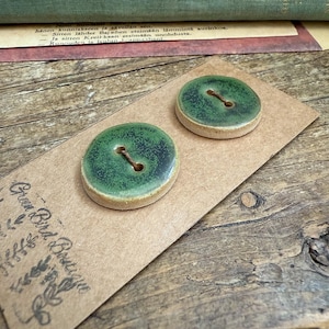 Op de afbeelding: Twee ronde, groene keramische knopen met een bruin knoopsgat, op een bruine kaart. De knopen hebben een gespikkelde groene glazuur en een beige rand. De kaart heeft de tekst "Green Bird Boutique" erop.