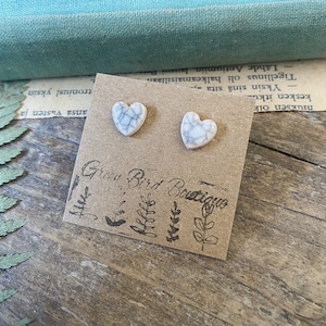 Könnte beinhalten: Ein Paar weiße herzförmige Ohrringe mit einer craquelé-Glasur. Die Ohrringe werden auf einer braunen Karte mit dem Text "Green Bird Boutique" präsentiert.