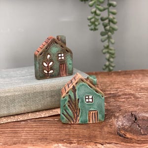 Könnte beinhalten: Zwei kleine Keramikhausfiguren mit einer texturierten grünen Glasur, braunen Dächern und handbemalten Details wie Fenstern, Türen und botanischen Motiven. Die Häuser sind mit einem rustikalen Finish und markanten Elementen gestaltet.
