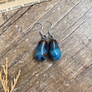 May include: A pair of silver teardrop earrings with blue and brown ceramic drops. The drops have a marbled effect.
