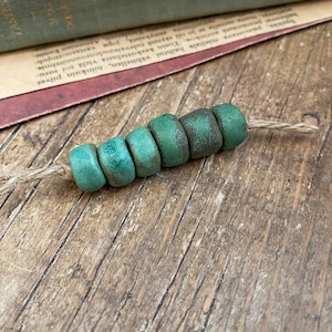 Pode incluir: Uma sequência de seis contas cilíndricas verde-turquesa com uma aparência rústica e envelhecida. As contas são enfiadas em um cordão de fio natural, sugerindo um componente de joalheria feito à mão. O fundo inclui livros antigos e uma superfície de madeira.