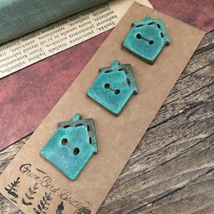 May include: Three ceramic house-shaped buttons in a teal green colour with brown stitching detail. The buttons are displayed on a brown card with the text "Green Bird Boutique".