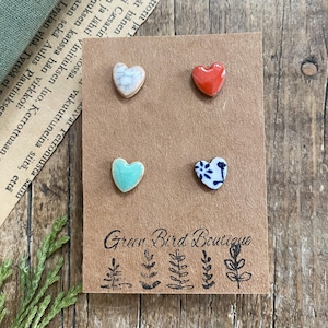 Może przedstawiać: Cztery kolczyki w kształcie serca w różnych kolorach i wzorach. Kolczyki są prezentowane na brązowej karcie z napisem "Green Bird Boutique" i ilustracjami botanicznymi.