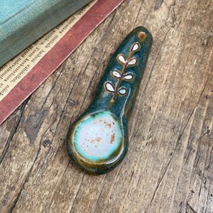 Può includere: Un piccolo cucchiaio in ceramica verde acqua con un motivo floreale bianco. Il cucchiaio ha una ciotola arrotondata e un manico lungo e stretto.