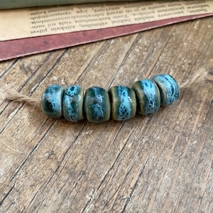 Può includere: Una fila di sei perline di ceramica con una glassa turchese e verde scuro, infilate su spago naturale. Le perline cilindriche hanno una fascia dorata al centro. Le perline sono esposte su una superficie di legno.