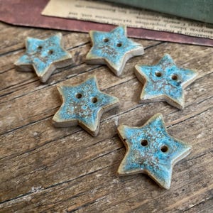 Può includere: Cinque bottoni a forma di stella con smalto turchese e marrone. Ogni bottone ha due fori per cucire. I bottoni sono disposti su una superficie di legno, mostrando il loro aspetto unico e fatto a mano.
