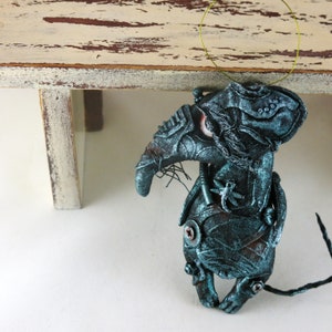 May include: A teal blue and silver steampunk-style rat figurine with a long, thin tail. The rat has a long, pointed nose and large, round eyes. The figurine is made of fabric and has a distressed, aged look.
