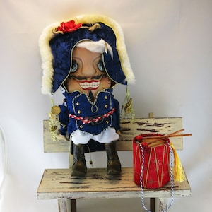 May include: A whimsical doll dressed as a military officer with a blue and gold uniform, a white hat with a red flower, and a white face with a wide smile. The doll is sitting on a wooden bench with a red and gold drum.