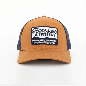 May include: A brown and navy blue trucker cap with a curved peak. The front panel displays a rectangular patch with the words "Tennessee Outfitters" and "Cookeville Tennessee Quality Goods For Quality Folks."