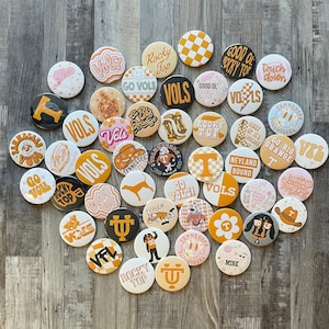 May include: A collection of circular pin-back buttons featuring various designs in orange, white, and black. The graphics include text such as VOLS, ROCKY TOP, GO VOLS, and NEYLAND BOUND, alongside illustrations of cowboy boots, hats, dogs, and checkerboard patterns.