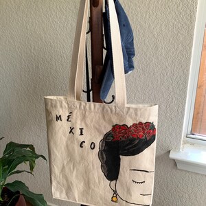 Op de afbeelding: Beige draagtas met lange cr&egrave;mekleurige banden. De tas heeft een zwart-rode illustratie van een vrouw met een bloemenkroon en het woord "M&Eacute;XICO" in zwarte letters. De tas hangt aan een houten kapstok.
