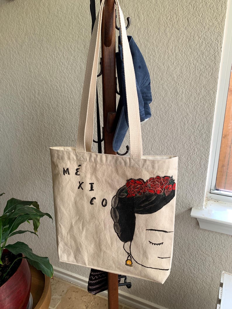 Op de afbeelding: Beige draagtas met lange cr&egrave;mekleurige banden. De tas heeft een zwart-rode illustratie van een vrouw met een bloemenkroon en het woord "M&Eacute;XICO" in zwarte letters. De tas hangt aan een houten kapstok.